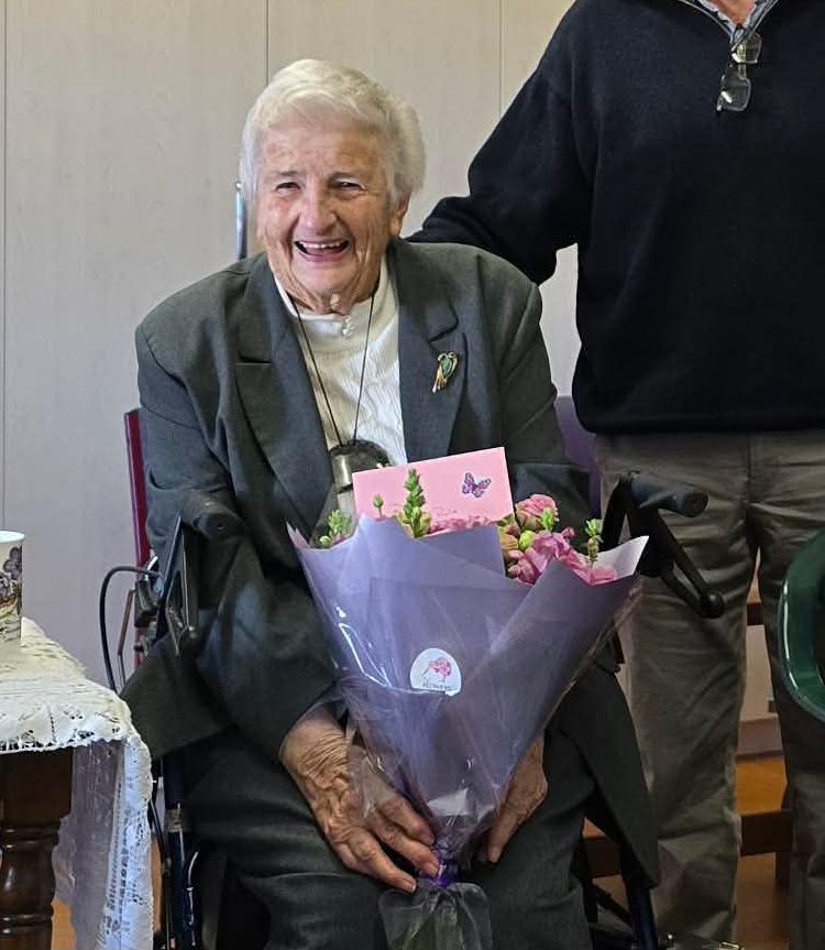 Topsy Rule QSM, seated and smiling, holding a bouquet of flowers